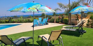 A poolside area with artificial grass, lounge chairs, and umbrellas. - By The Beach - Emerald Seaview Villa - Stunning Seaview Stay (Protaras)