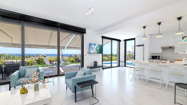 A modern living room with a large window, a sofa, a coffee table, and a dining area with bar stools.