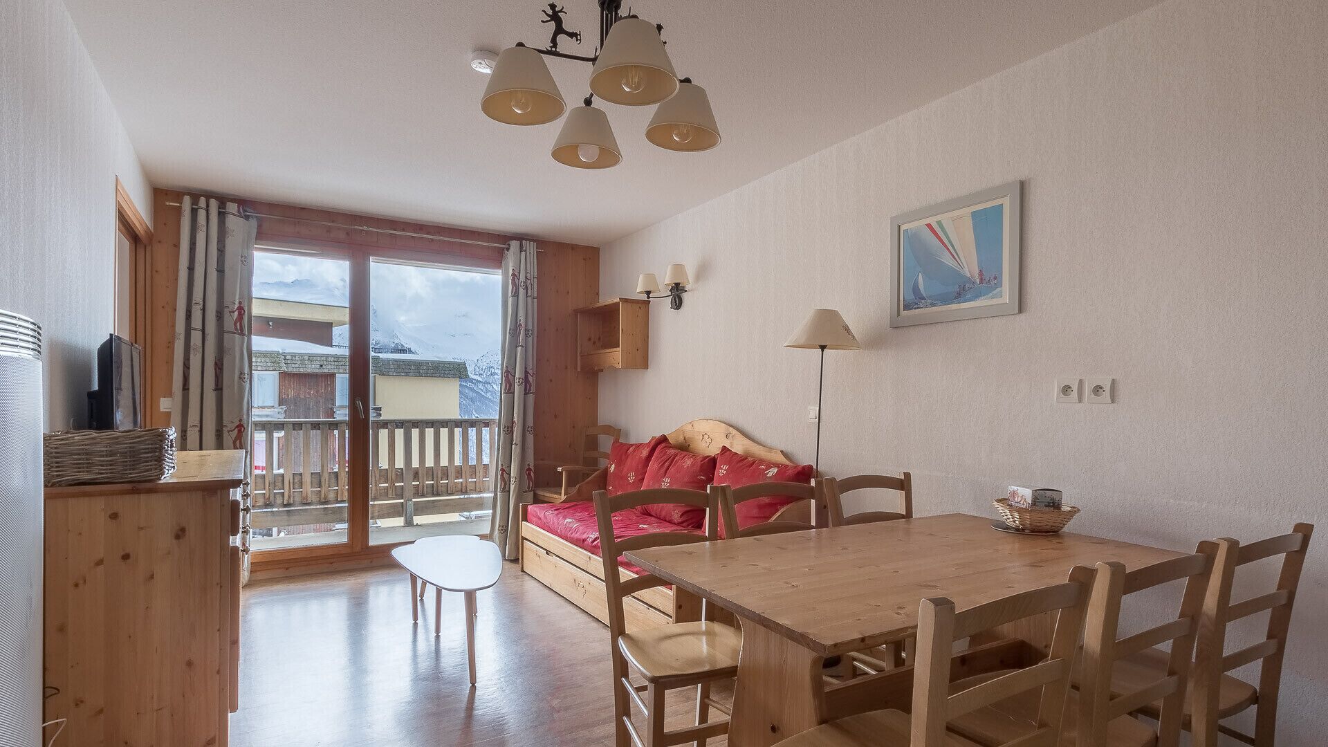 Ein gemütliches Zimmer mit einem Holzesstisch, einem roten Sofa und Blick auf Berge vom Balkon.