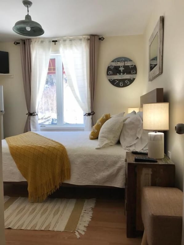 Un dormitorio con cama, lámpara de noche, un reloj en la pared y ventana con cortinas.