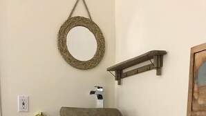 A rustic bathroom with a wooden cabinet, a hanging towel, a woven mirror, and a small shelf. - auberge maison gagne (Tadoussac)