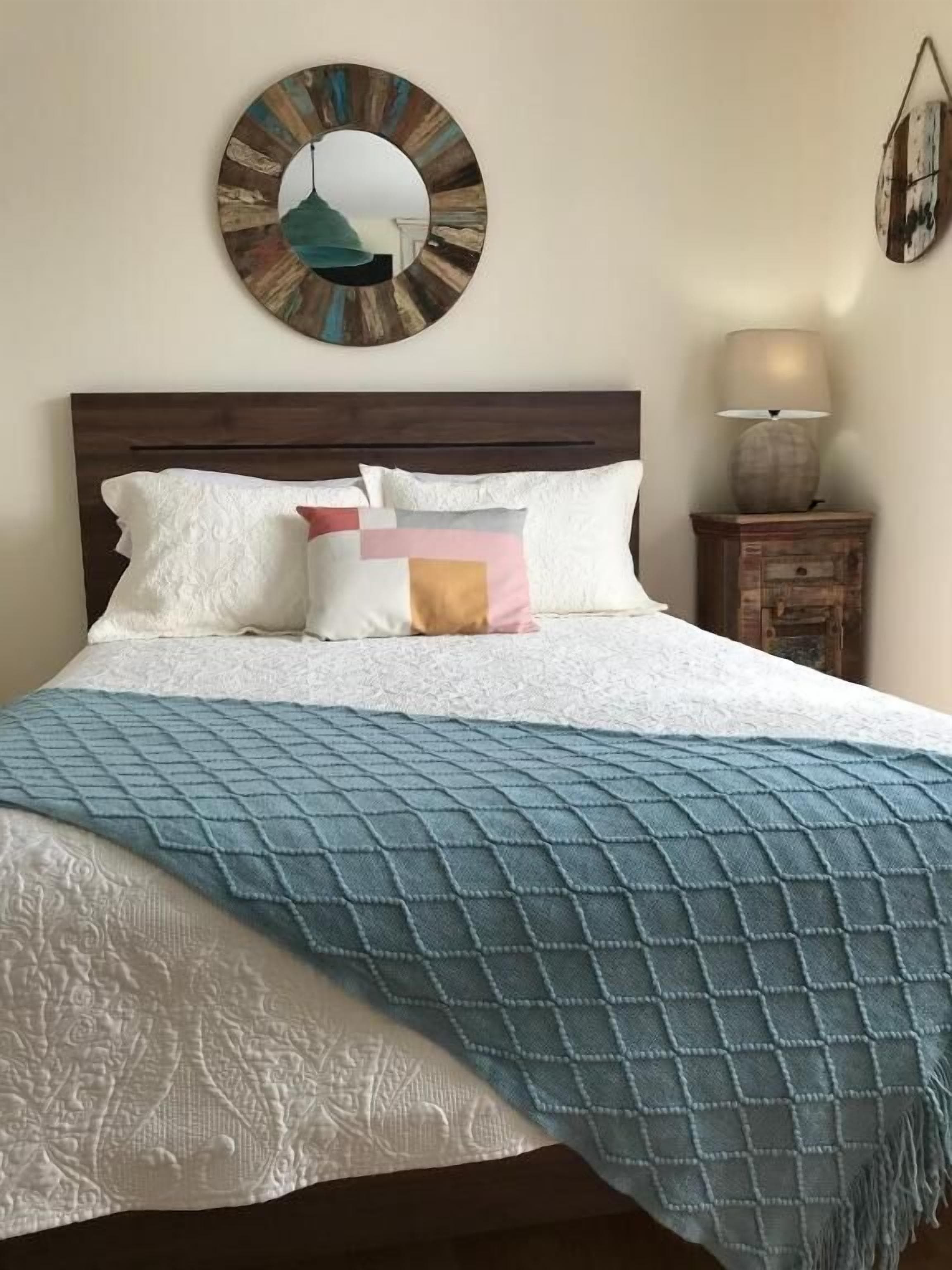 A neatly made bed with a quilted blue blanket, white pillows, and a wooden headboard. A round mirror with a rustic frame hangs on the wall above the bed.