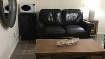 Ett vardagsrum med en svart lädersoffa, ett soffbord i trä, en väggklocka, en mikrovågsugn och en stor, rund spegel.