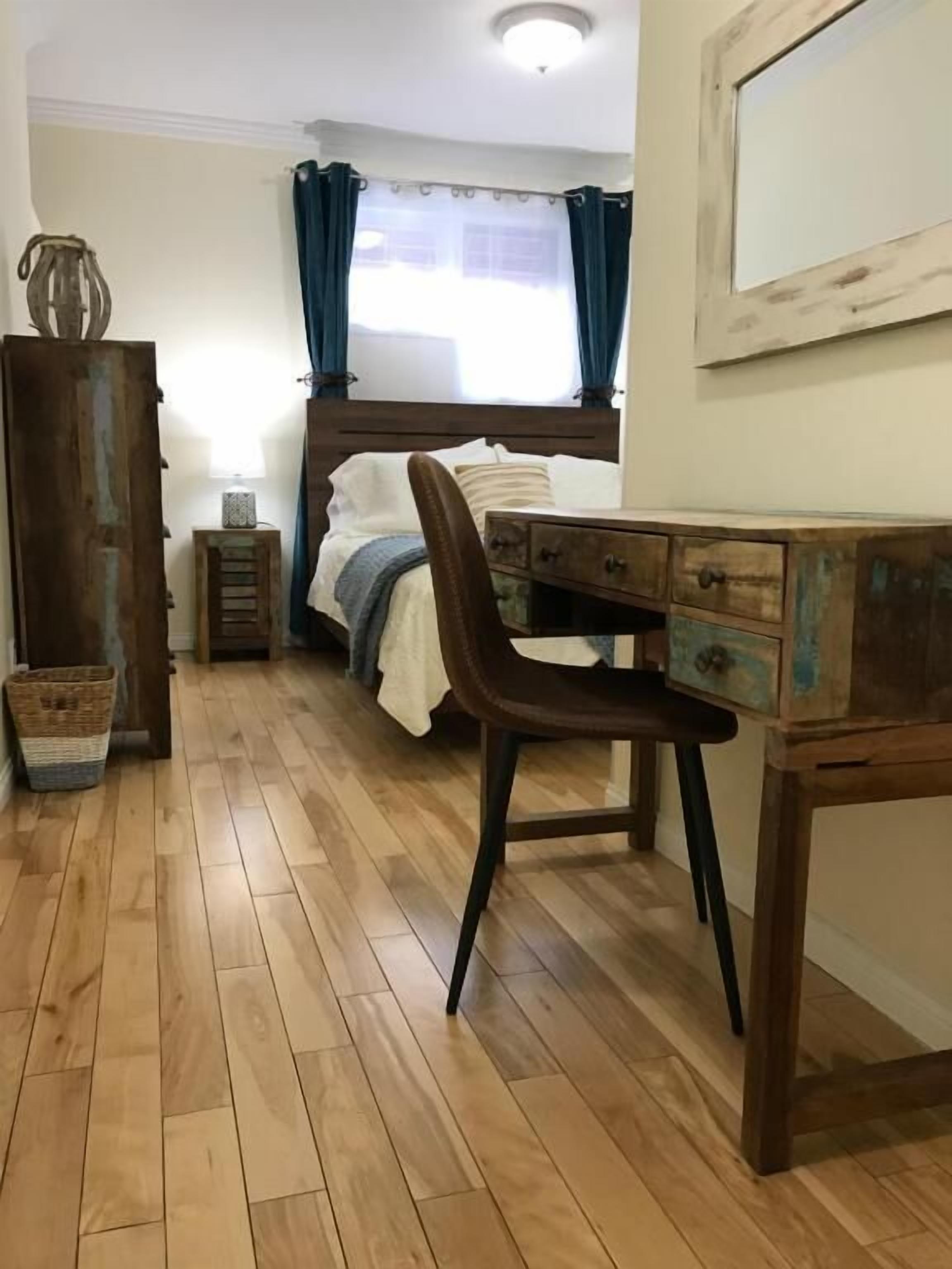 Een slaapkamer met een houten bureau, een bed met hoofdeinde, een nachtkastje, een lamp en een raam met gordijnen.