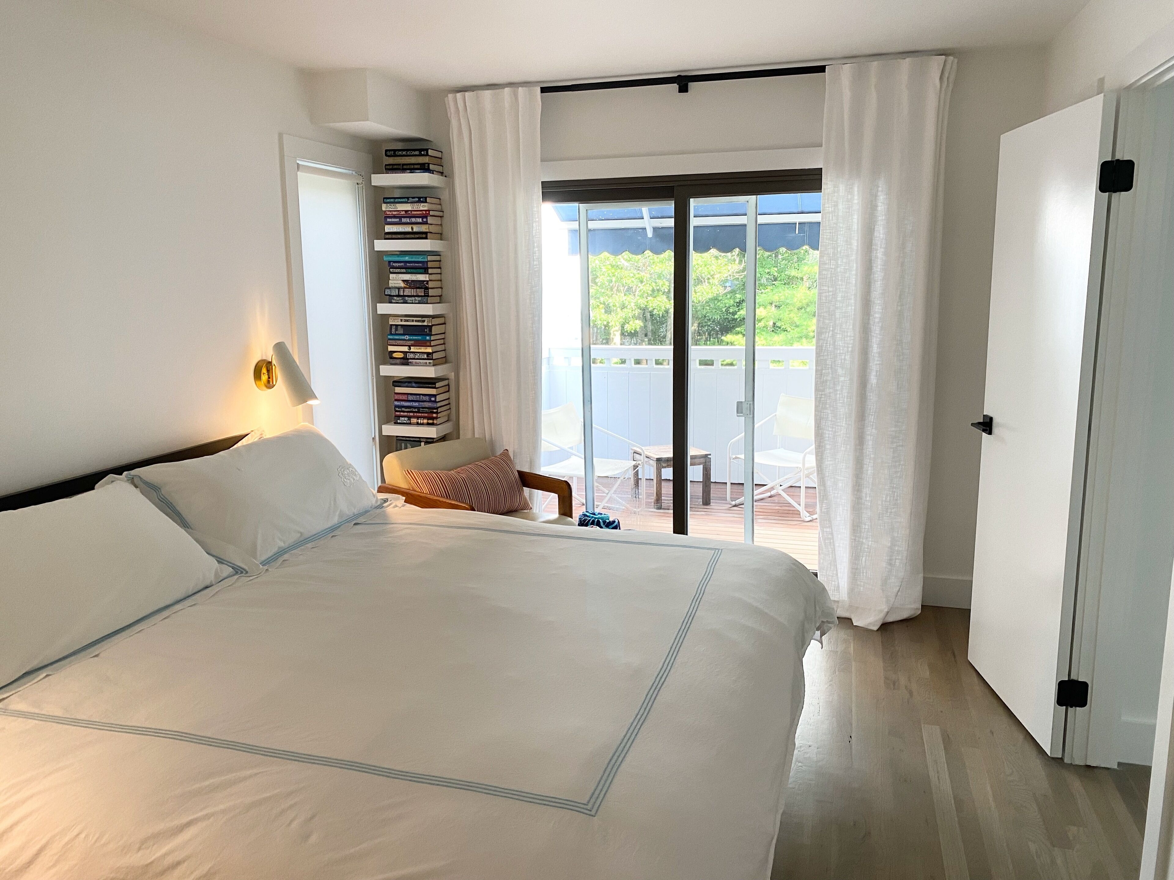 A bedroom with a large bed, a bedside table with a lamp, a bookshelf, and a sliding glass door leading to an outdoor patio with a table and chairs.