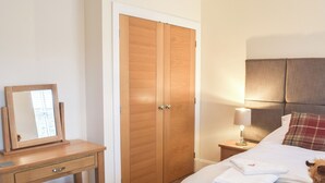 Quarto de hotel com armário de madeira, mesa de cabeceira com abajur, cama com manta branca e travesseiro vermelho, e um pequeno banco de madeira.