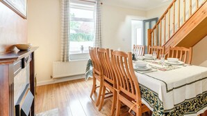 Ein Essbereich mit einem Holztisch, gedeckt für vier Personen, flankiert von Holzstühlen. Ein Fenster mit Vorhängen ist sichtbar, und eine Treppe führt zu einer oberen Ebene.