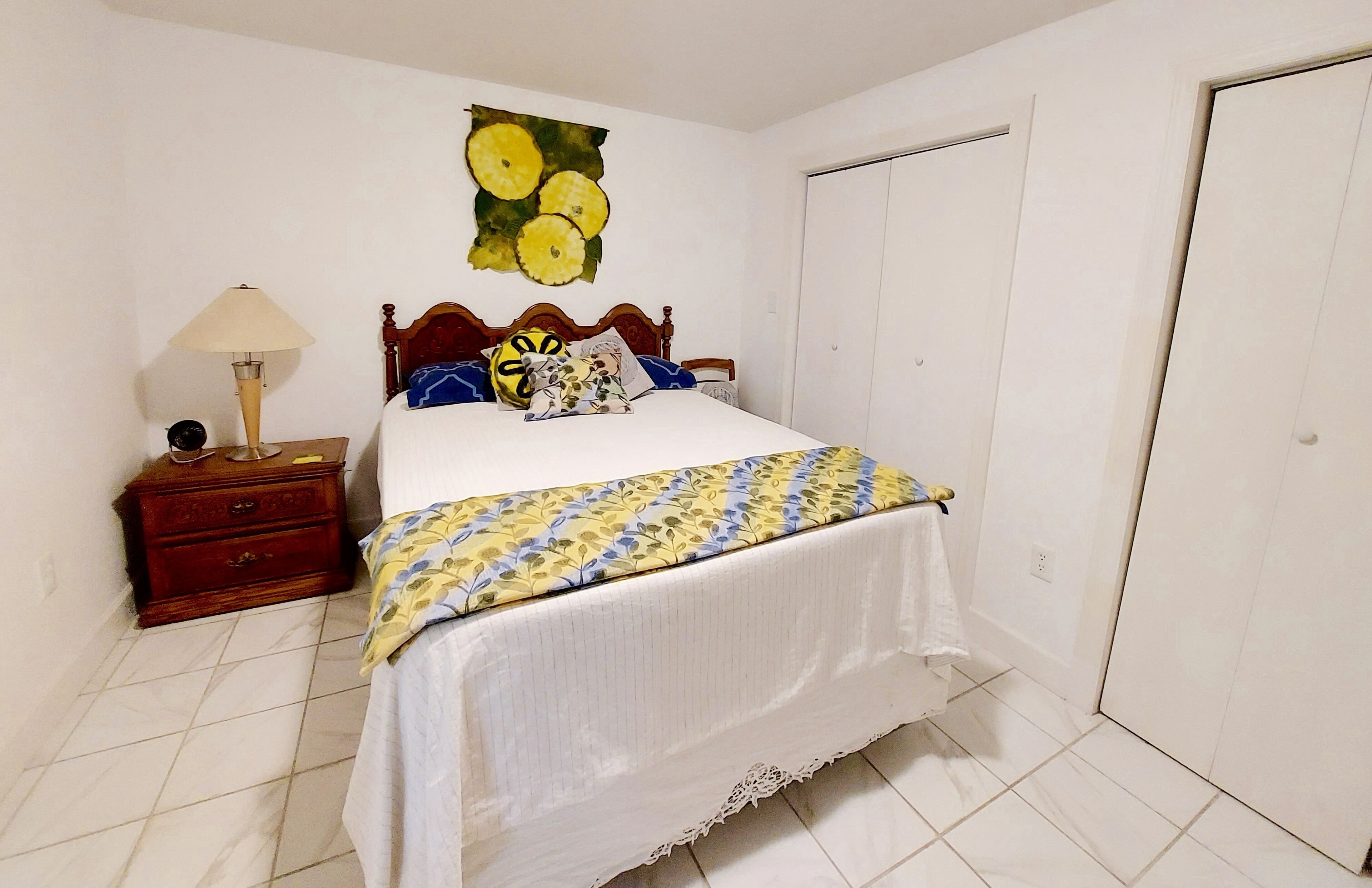 A bedroom with a bed, a nightstand, a lamp, a wooden headboard, a wall painting, and a closet.