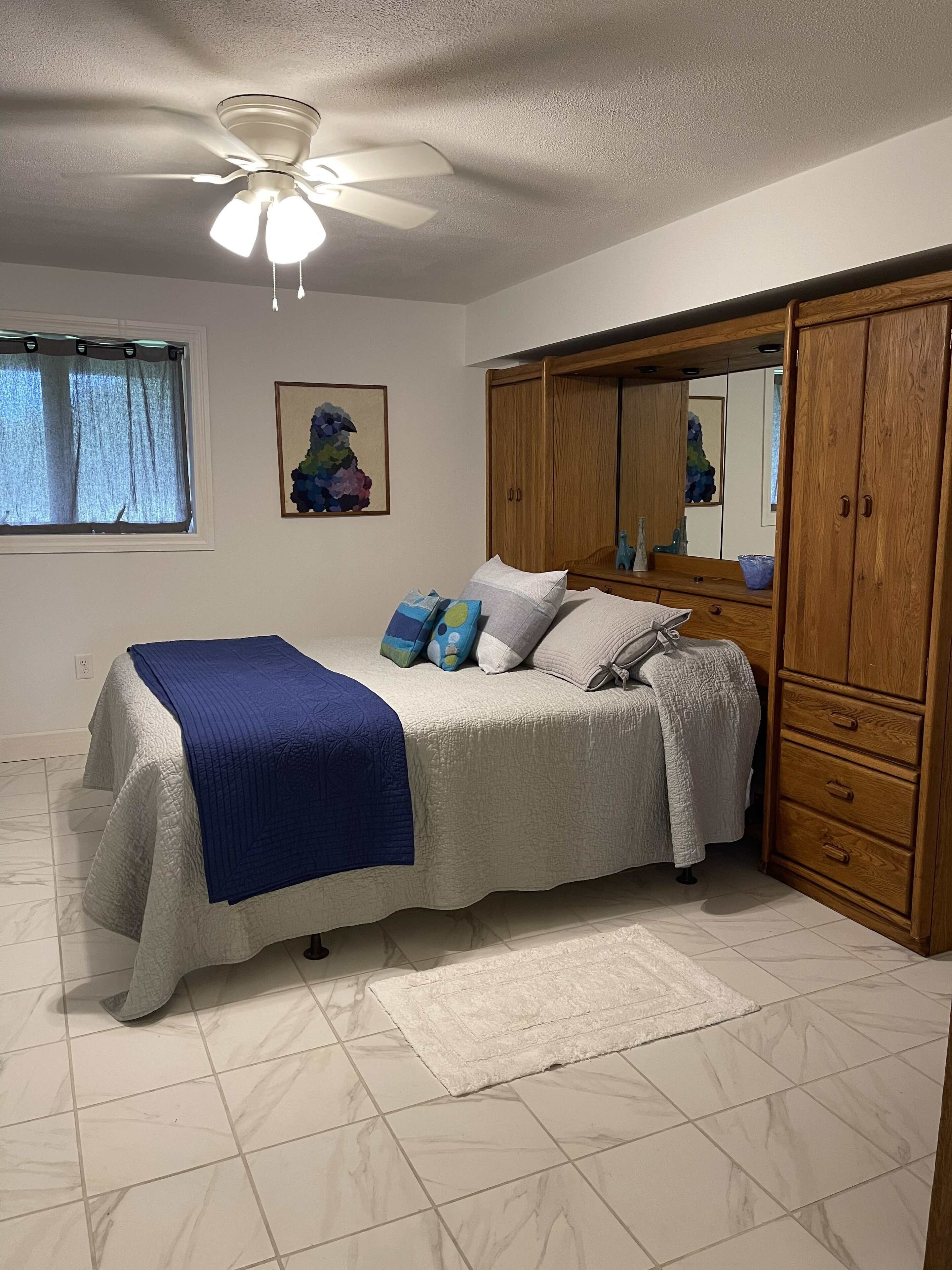 Quarto com cama, guarda-roupa, ventoinha no teto, janela com cortinas e uma obra de arte na parede.