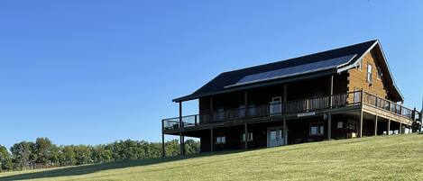 A two-story lodge with a balcony, solar panels, and a grassy hill in the foreground.