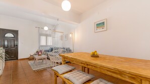 A dining area with a wooden table and benches, a white chair, a small side table with a vase of flowers, and a framed picture on the wall. - Sweet 2-Bedroom Townhouse (Lagos)