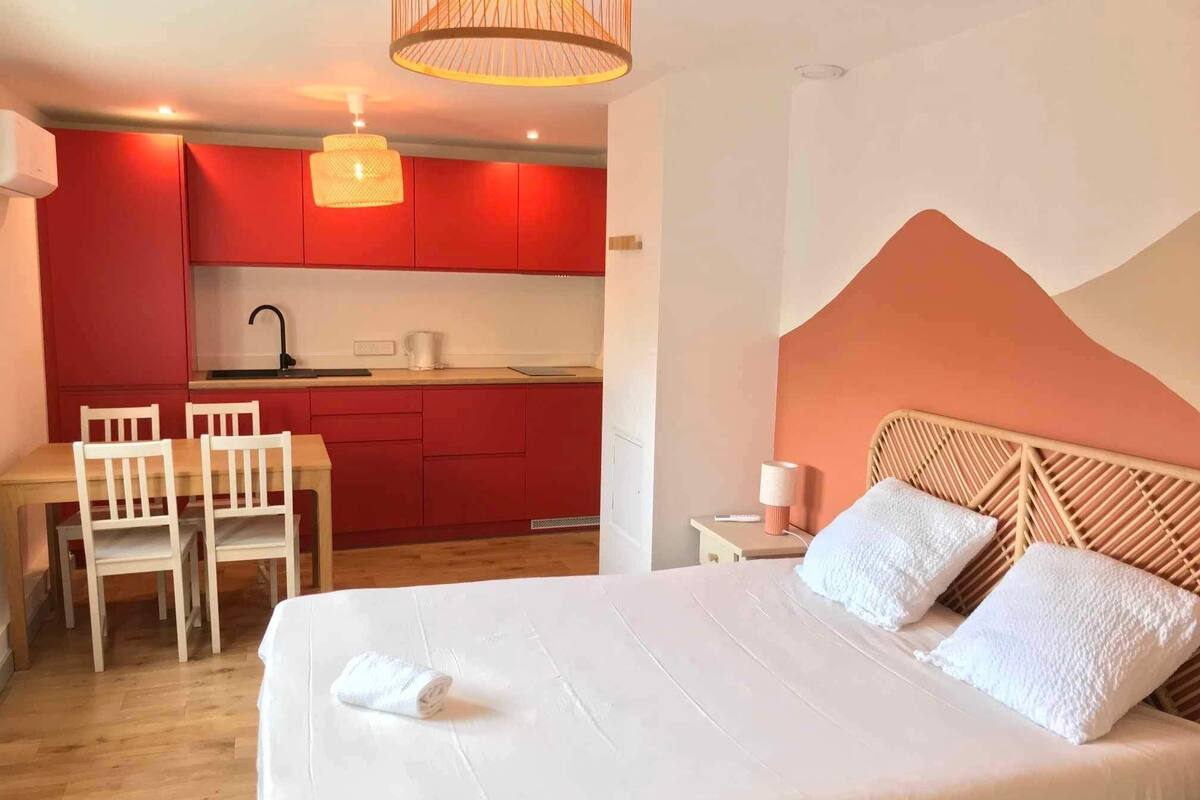 Une chambre à coucher avec un lit, une table de chevet, une lampe et une vue sur une cuisine rouge.