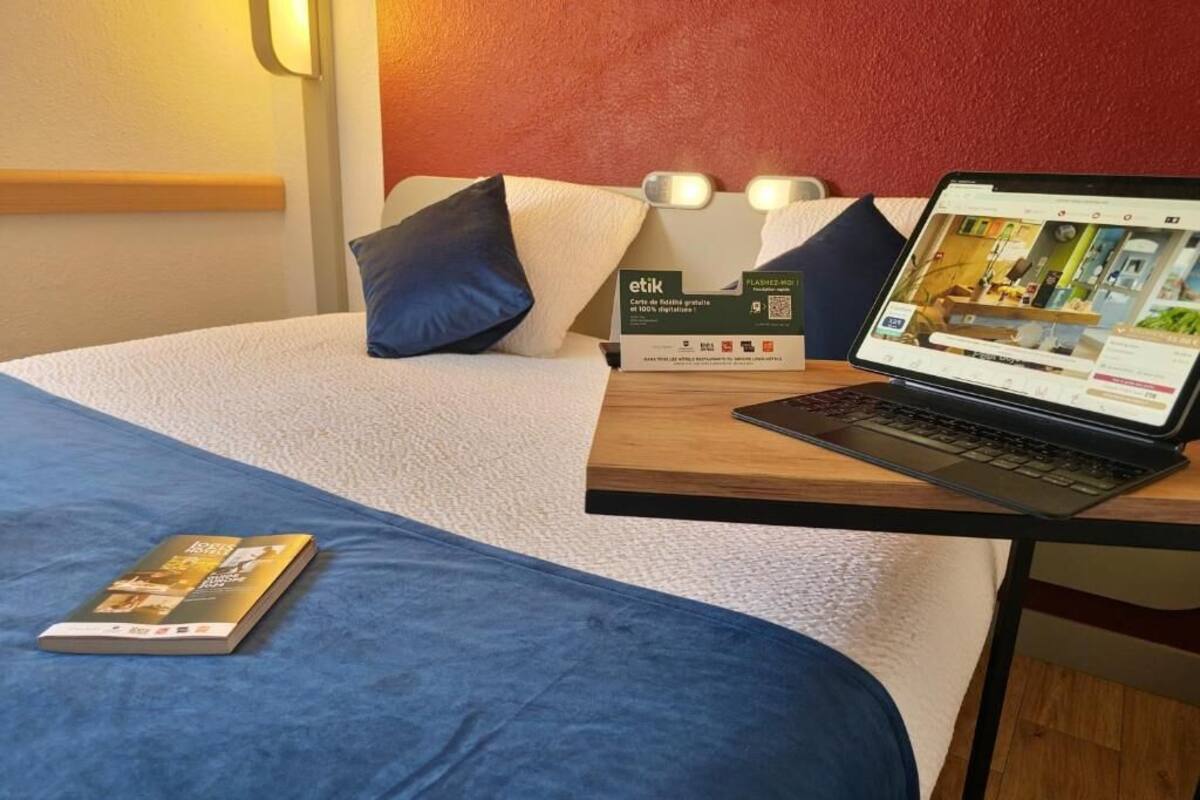 Un lit bien fait, avec une couverture bleue et des oreillers, un ordinateur portable posé sur une table de chevet en bois, et une brochure sur le lit.
