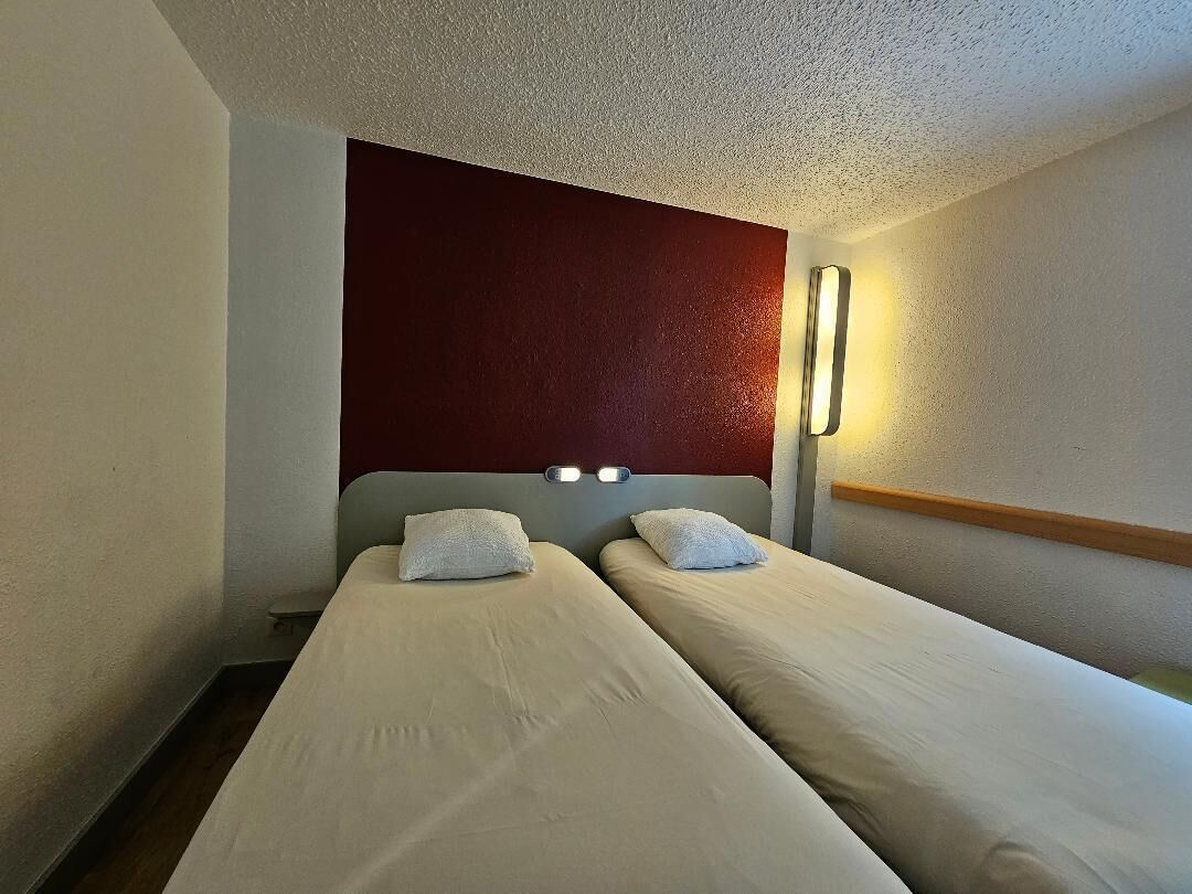 Une chambre d’hôtel avec deux lits simples, un mur d’accent rouge et un luminaire fixé au mur.
