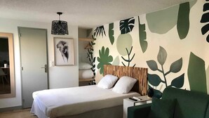 Een slaapkamer met een bed, een groene bank, een houten hoofdbord, een muur met bladmotieven, een hanglamp en een lijst met een afbeelding.