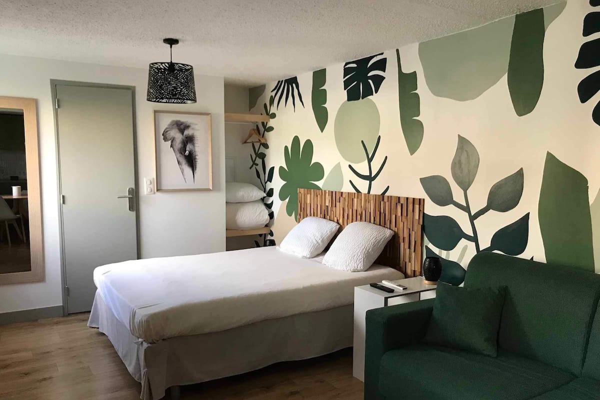 Une chambre avec un lit, un canapé vert, une tête de lit en bois, un mur orné de motifs de feuilles, un luminaire suspendu et un tableau encadré.