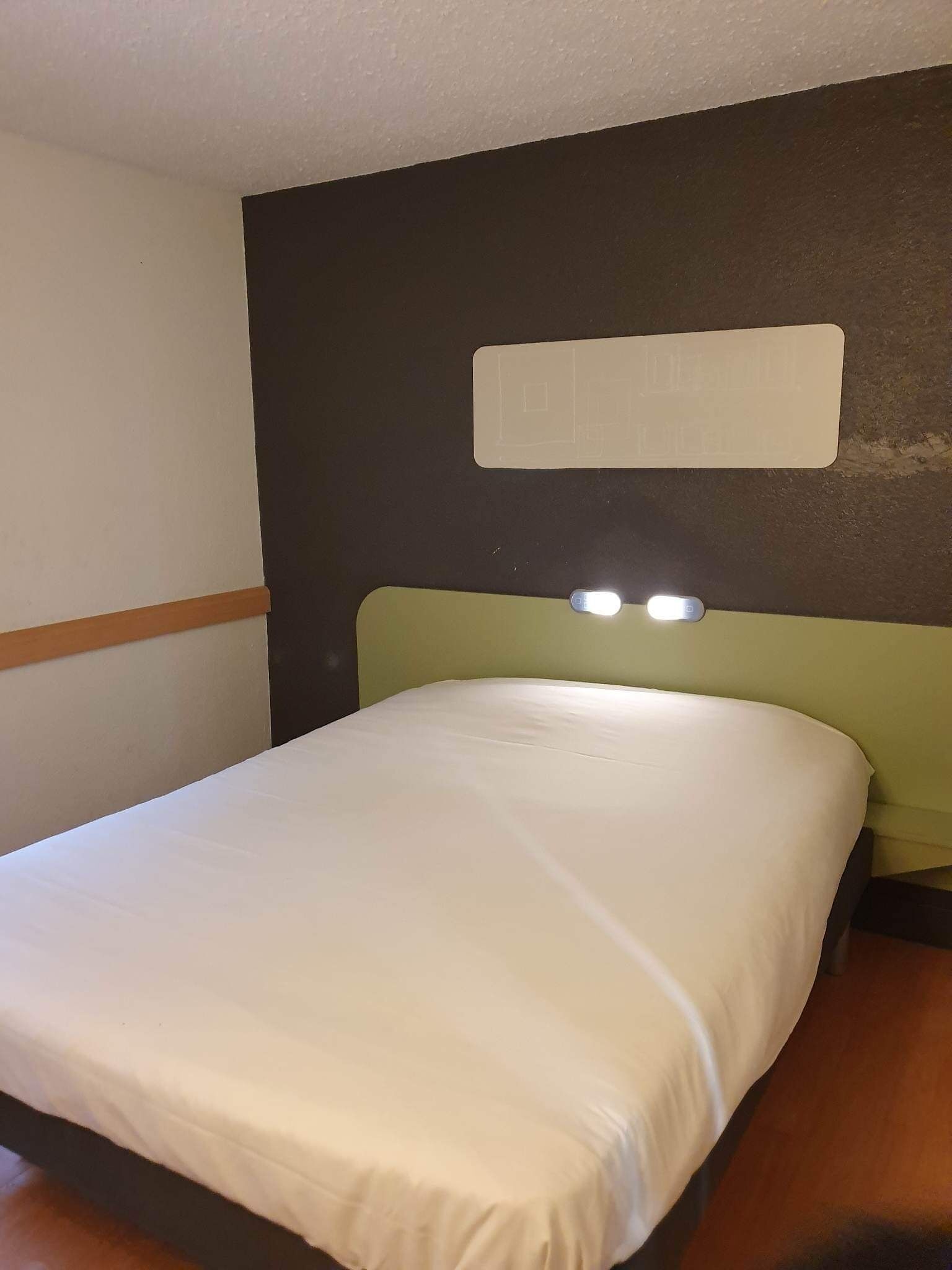 Een hotelkamer met een bed, een hoofdeinde met verlichting en een bedieningspaneel aan de muur.