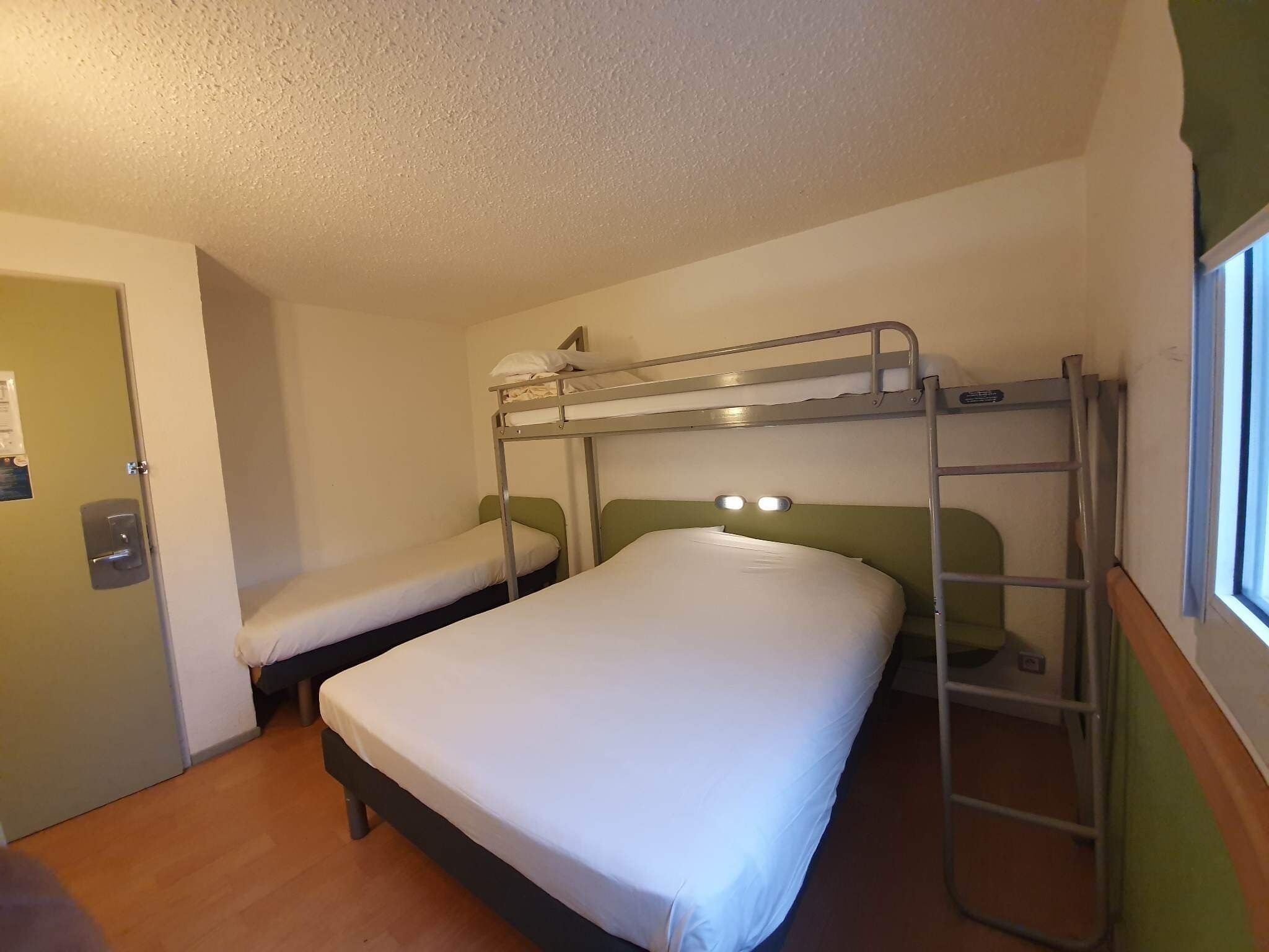 Een slaapkamer met een stapelbed, een enkel bed en een bed er bovenop. Een raam aan de rechterkant en een deur aan de linkerkant.