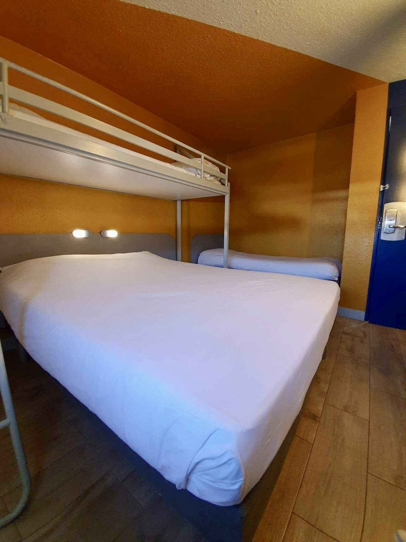 Habitación con literas, suelo de madera, colcha blanca y puerta con cerradura.
