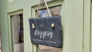 Une pancarte noire, sur laquelle est écrit « The Piggey Carlingcott, Somerset », est accrochée à une porte verte.