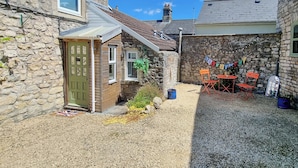 Une maison en pierre avec une porte verte, un petit patio avec une table et des chaises, et une allée gravillonnée.
