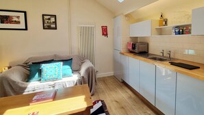 Une cuisine de taille réduite, dotée d’un four encastré, d’un évier et d’un plan de travail. Une fenêtre se trouve au-dessus de l’évier, et un radiateur est visible sur le mur.