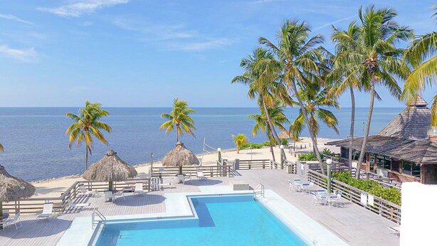 Zona de piscina en un resort con vista al océano, palmeras y estructuras de techo de paja.