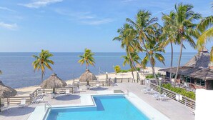 A resort pool area with a view of the ocean, palm trees, and thatched-roof structures. - Barefoot Bliss: Ground Floor Condo with Den & Dual Balconies (Islamorada)