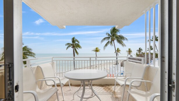 A balcony with a white table and chairs, overlooking a beach with palm trees and a clear sky. - Second Floor Ocean View Studio – Gorgeous Waterfront Escape (Islamorada)