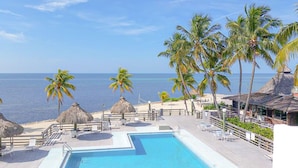 Zwembadgebied van een resort met uitzicht op de oceaan, palmbomen en constructies met rieten daken.