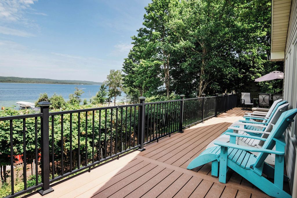 A wooden deck with a black railing, overlooking a lake. There are turquoise Adirondack chairs and a table with a canopy.