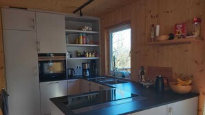Uma cozinha moderna com fogão, forno embutido e pia. Há uma janela que proporciona luz natural e uma prateleira com diversos utensílios de cozinha.