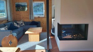 Um quarto acolhedor em estilo cabana, com interior de madeira, um sofá, uma mesa de centro, uma lareira e uma grande janela com vista para uma paisagem montanhosa.