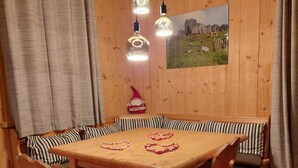 Uma sala de jantar de madeira com uma mesa, cadeiras e um banco. Há cortinas, um quadro na parede e uma almofada vermelha.