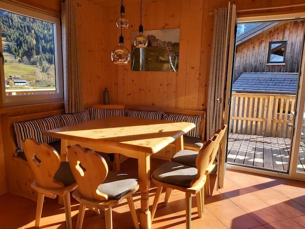 A wooden interior with a round table and benches, a large window with a view of a forest, and a door leading to an outdoor area.