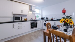 Moderni keittiö, jossa on valkoiset kaapit, sisäänrakennettu uuni, mikroaaltouuni ja pesukone. Keittiössä on ruokapöytä, jonka päällä on maljakko auringonkukkia ja pullo viiniä.