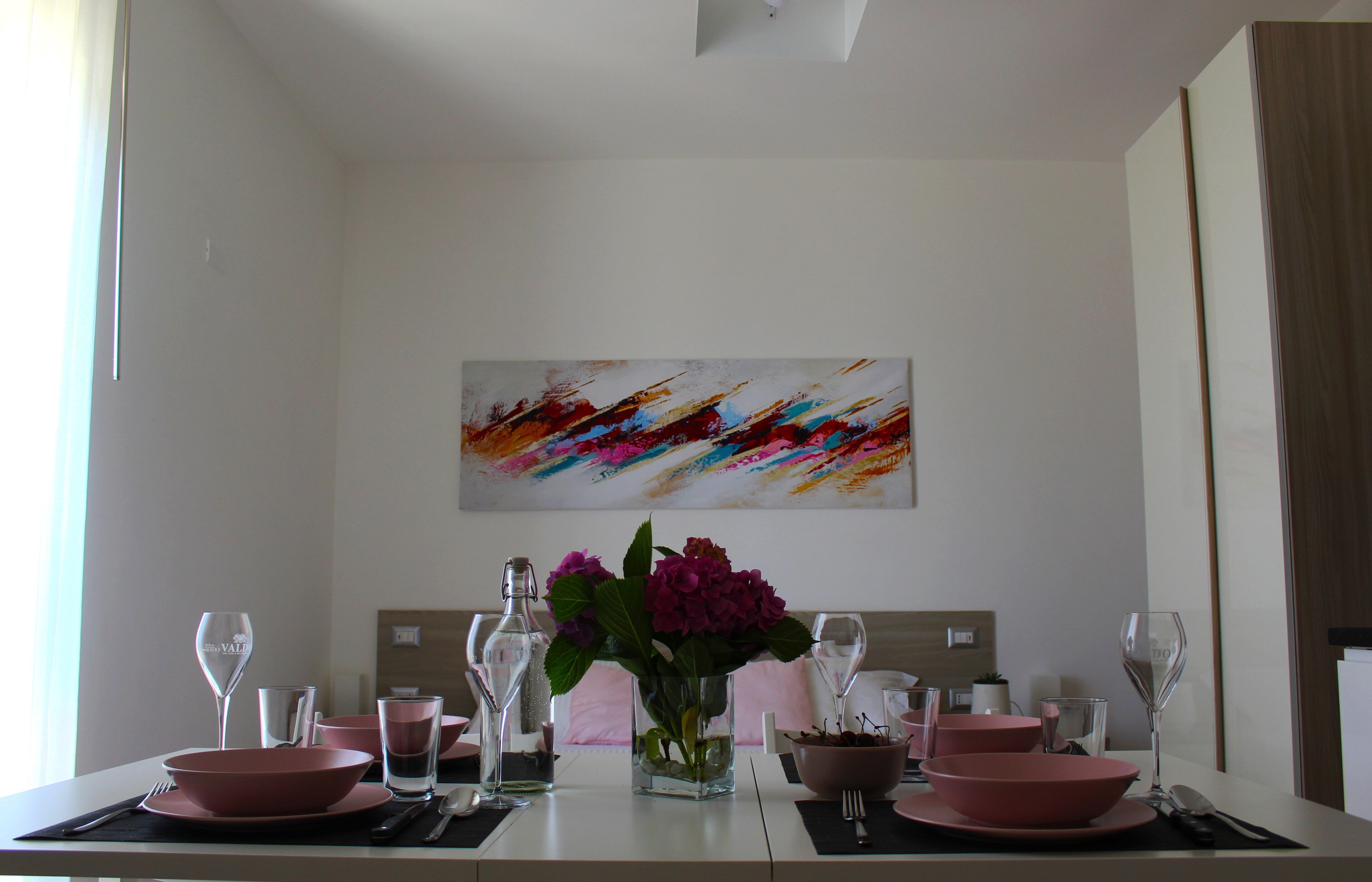 Une table à manger dressée avec des bols, des assiettes et des couverts roses, un vase rempli de fleurs et un tableau abstrait aux couleurs vives accroché au mur.