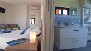 Ett modernt hotellrum med en säng, ett sängbord, en lampa och ett badrum med handfat, spegel och handduksställ.