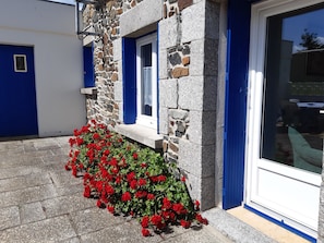 Un edificio de piedra con puertas y ventanas azules, un pequeño porche con flores rojas y un cielo despejado.