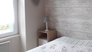 A bedroom with a bed, a nightstand, a wall-mounted lamp, a window, and a decorative heart on the wall. - Maison Bord de mer avec Jardin clos 3 CH/7 Personnes Plage à 550m (HILLION)