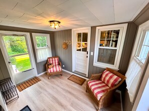 A cozy room with a wooden floor, two wicker chairs with plaid cushions, a white door, and a window overlooking a green lawn.