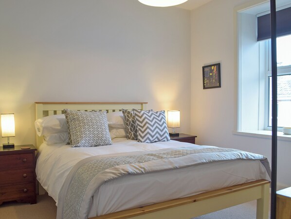 A neatly made bed with patterned pillows, a bedside lamp, and a window with blinds. - 5 bedroom accommodation in New Quay (New Quay)