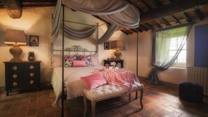 Ein Himmelbett mit floraler Bettwäsche, ein Nachttisch mit Lampe und eine Bank.