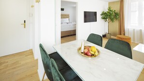Habitación de hotel moderna con una mesa de comedor de mármol, sillas tapizadas en verde, un frutero, un televisor de pantalla plana y una planta en maceta.