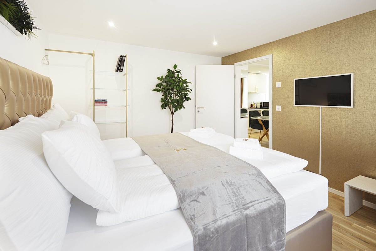 A modern hotel room with a large bed, a bedside table, a TV, a plant, and a view of the kitchen through an open door.