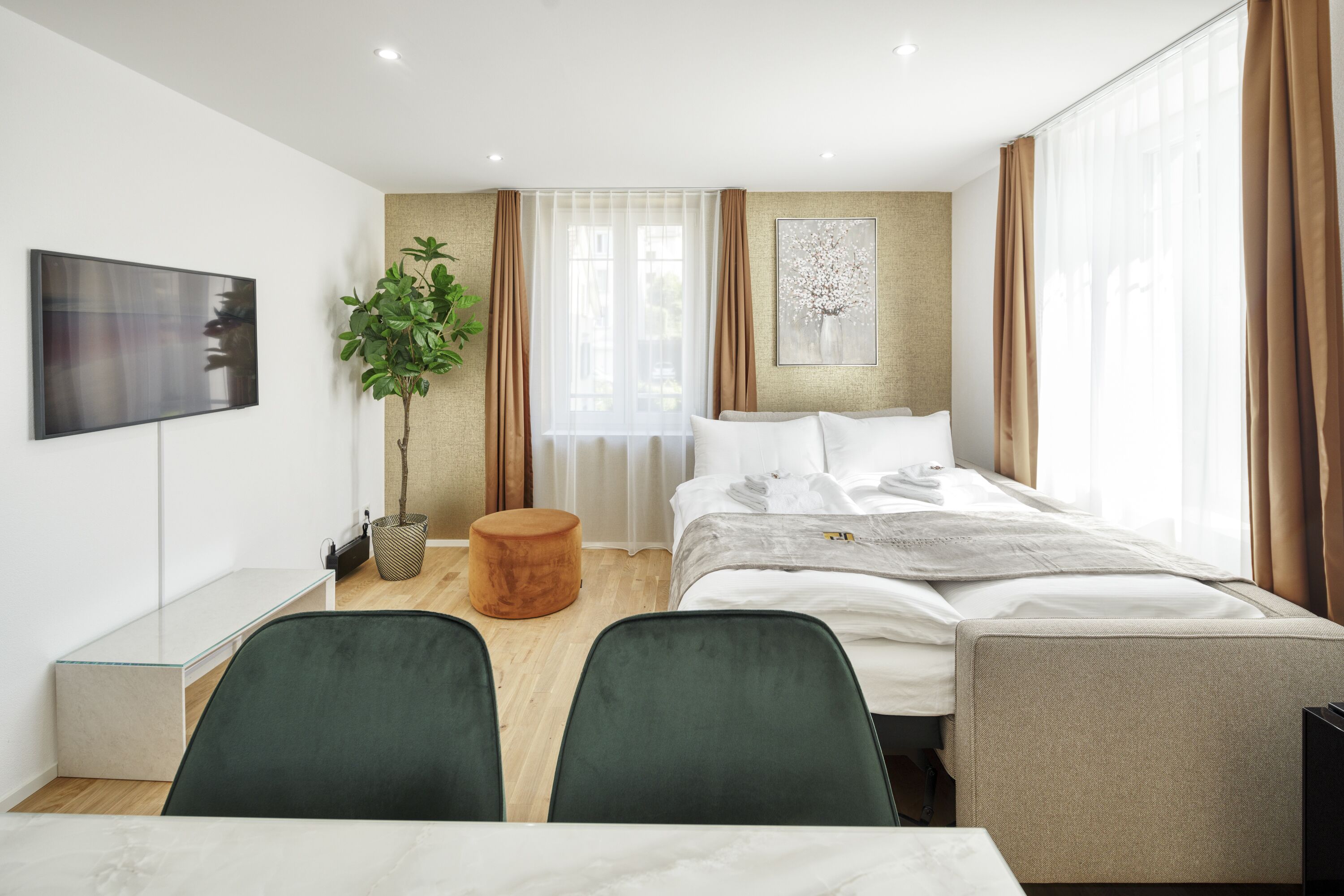 A modern hotel room with a bed, two chairs, a TV, a plant, and a window with curtains.