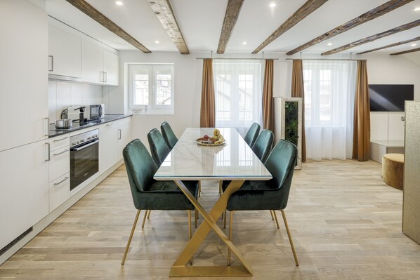 Una cocina moderna con armarios blancos, horno empotrado y comedor con mesa de mármol y sillas tapizadas en verde.