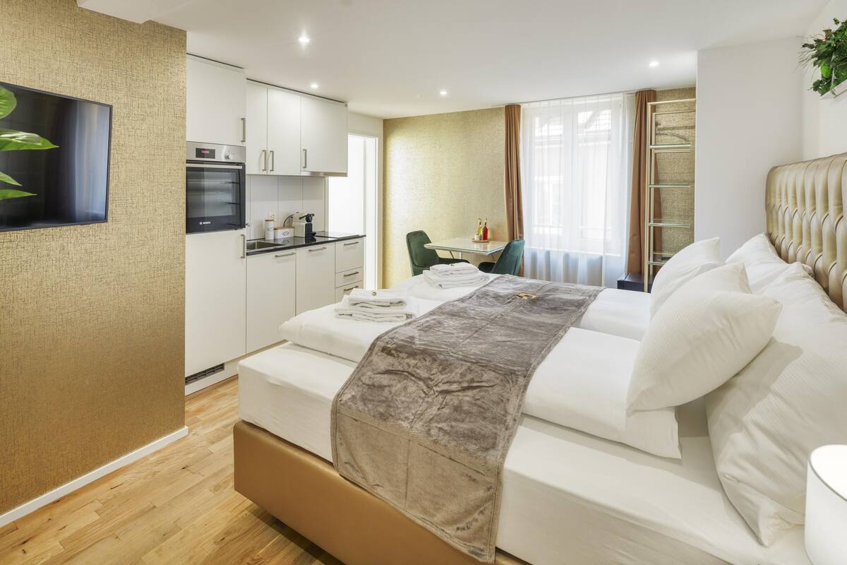 A modern hotel room with a bed, a small kitchenette, a dining area, and a large window with curtains.