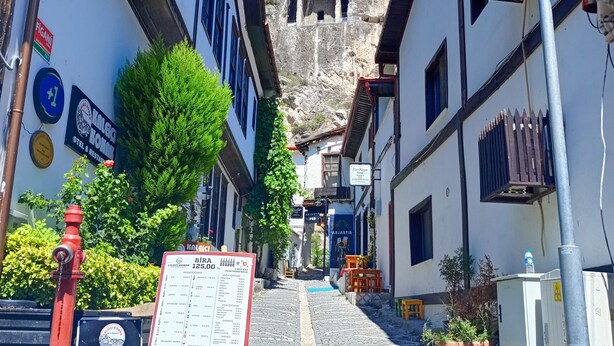 Uma rua de paralelepípedos estreita, com edifícios em ambos os lados, uma placa de sinalização e uma grande formação rochosa ao fundo.