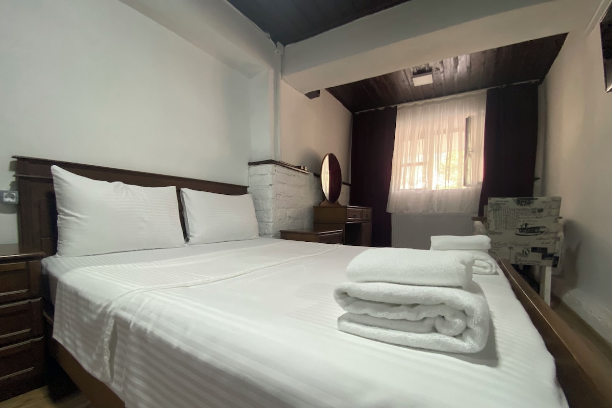 A neatly made bed with white linens, a wooden headboard, a bedside table with a mirror, and a window with curtains.